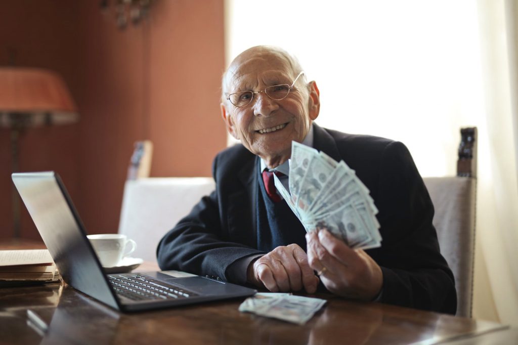 older white man seated in front of a laptop with a bunch of paper currency